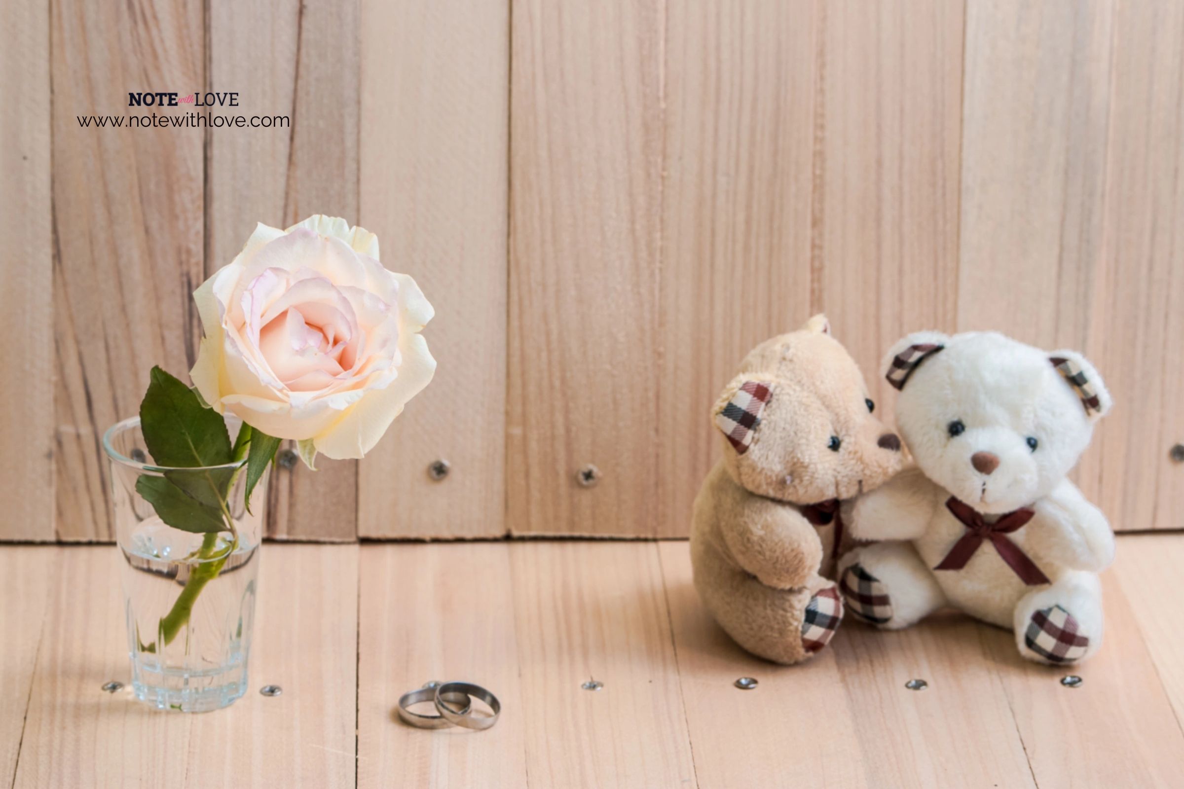 A white and a brown teddy bear hugging each other. A rose is kept in water in a glass beside these.